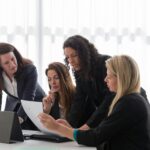 Four businesswomen engaged in discussion over documents in a contemporary office setting.