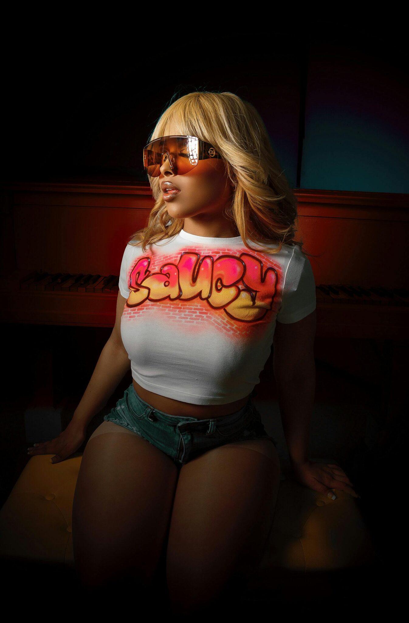 Stylish woman in crop top and denim shorts poses dramatically indoors with sunglasses.