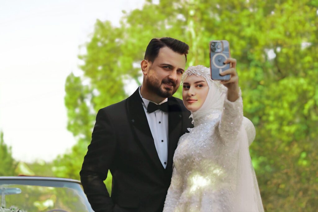 Bride and groom in elegant attire taking a selfie outdoors with lush greenery in the background.